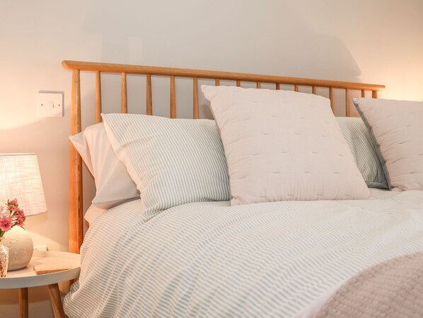 Una cama bien hecha con una colcha a rayas y almohadas, una mesita de noche con una lámpara y un jarrón con flores, y un cabecero de madera.