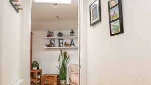 En hall som leder in till ett vardagsrum med trägolv, ett litet bord med en växt och en soffa. Det finns hyllor med dekorativa föremål och en skylt med texten "HAV".