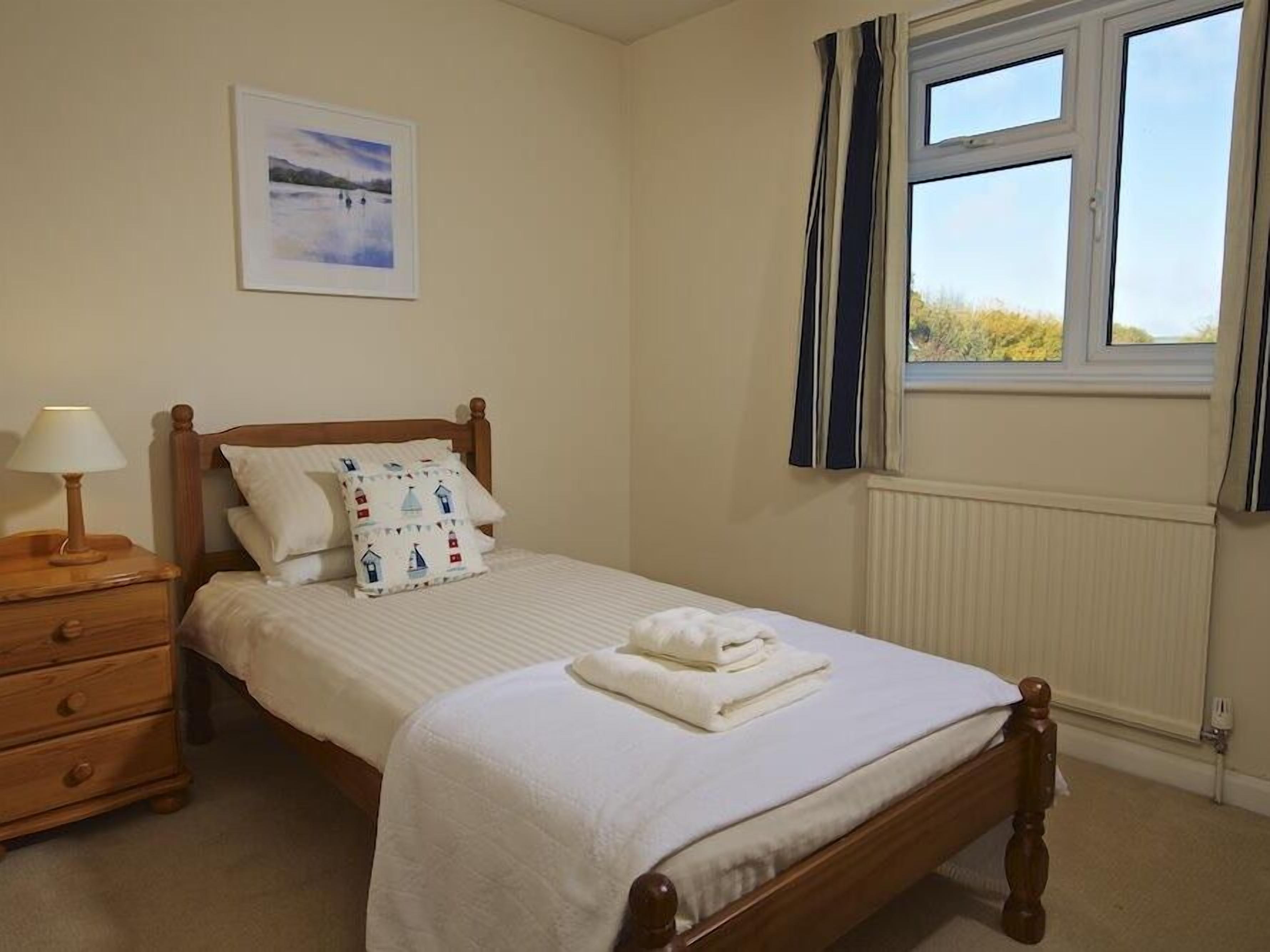 Een netjes opgemaakt bed met een witte dekbedovertrek en kussens, een nachtkastje met een lamp, en een raam met gordijnen.