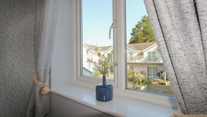 Una ventana con vista a una zona residencial, un jarrón azul con flores y un adorno de cuerda.