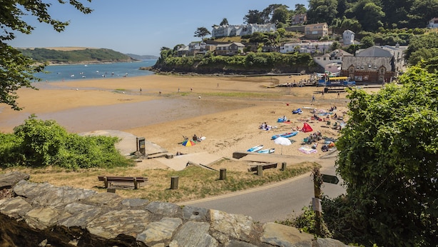Una playa costera con escalones de piedra que bajan hacia la orilla, zona de arena con sombrillas y gente disfrutando, y al fondo casas y vegetación.