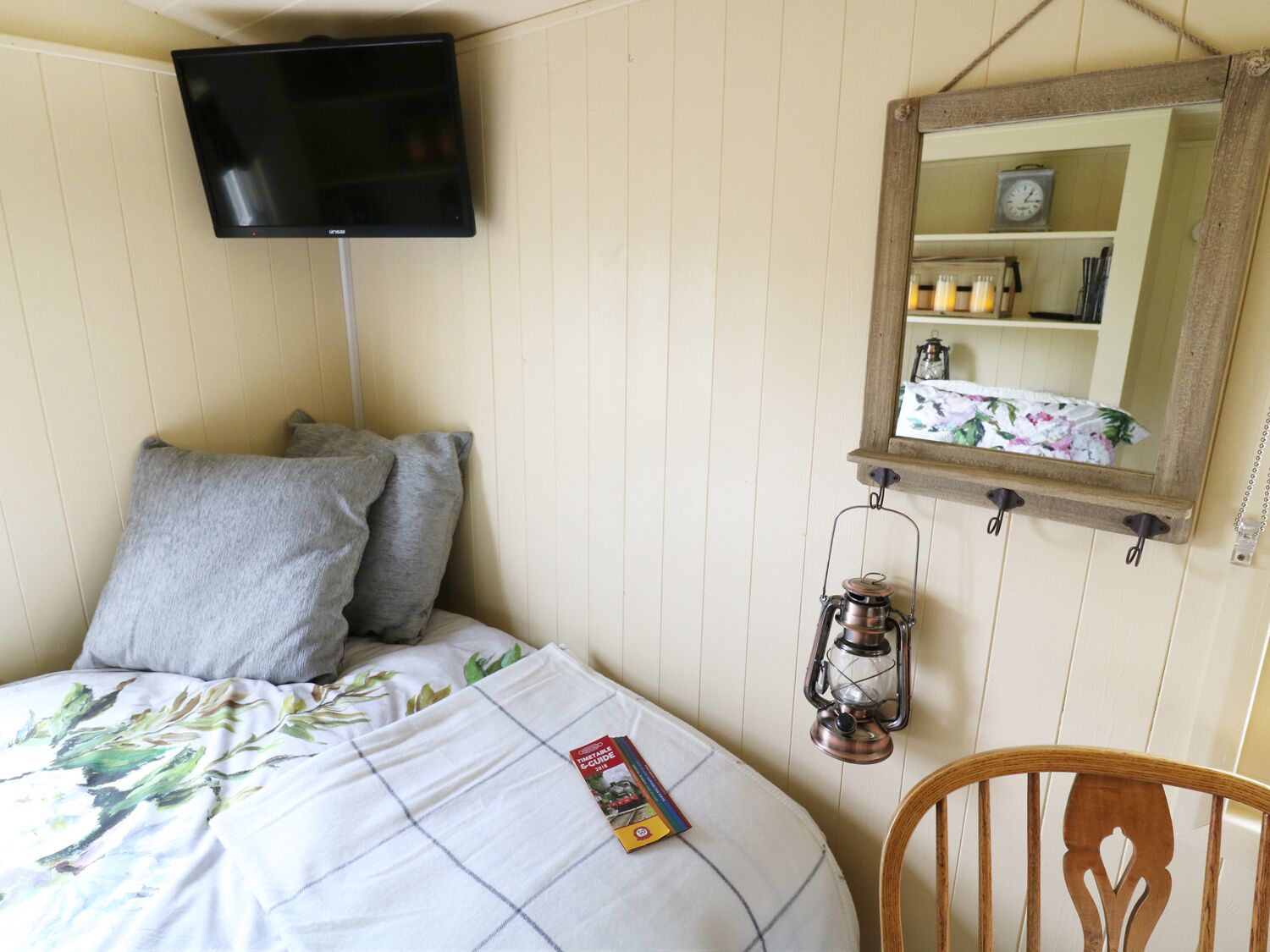 Un dormitorio con cama, almohadas, televisión, espejo, farol y una silla.