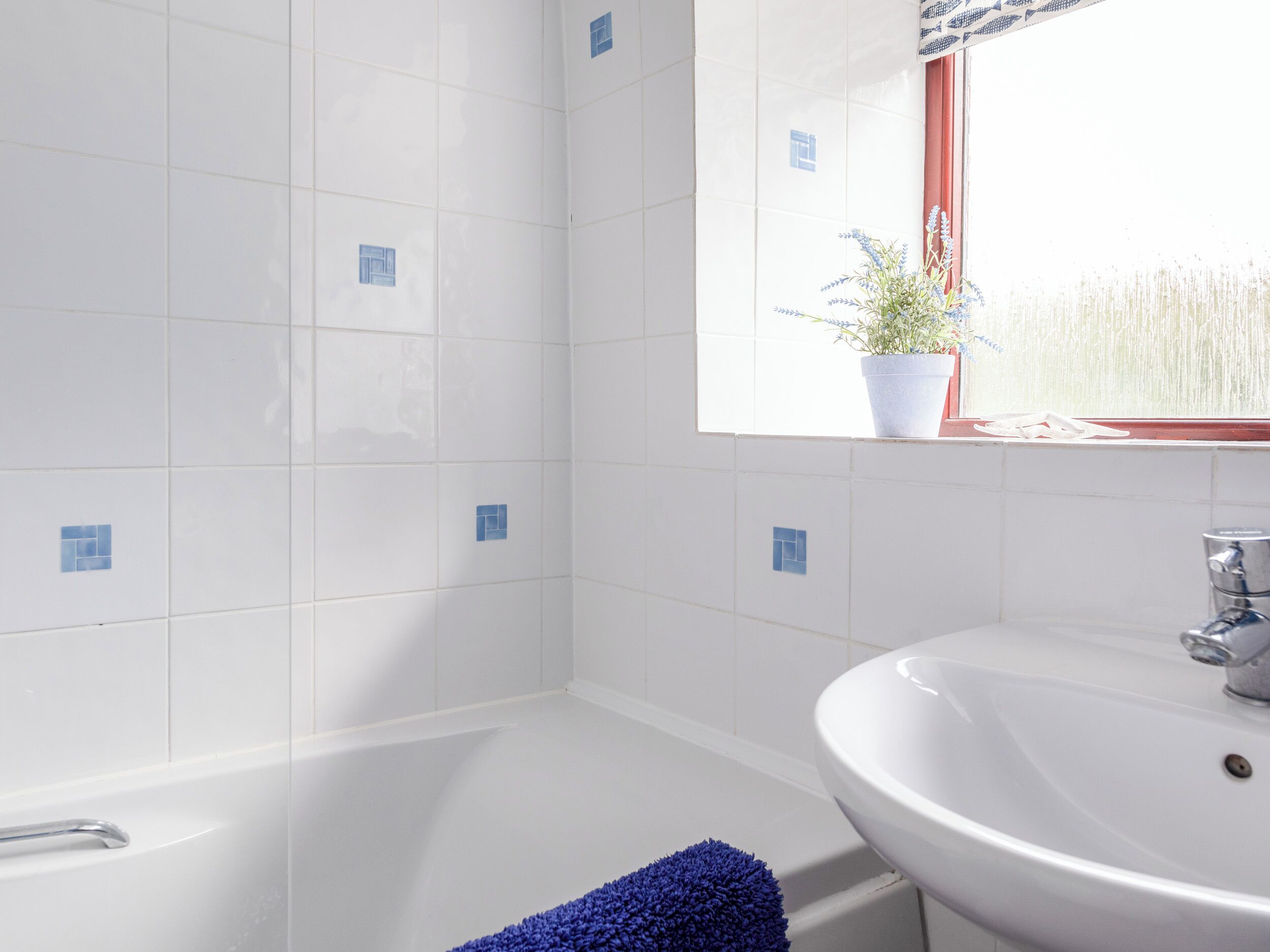 A bathroom with white tiles, a white sink, a window with a plant, and a blue towel on the floor.