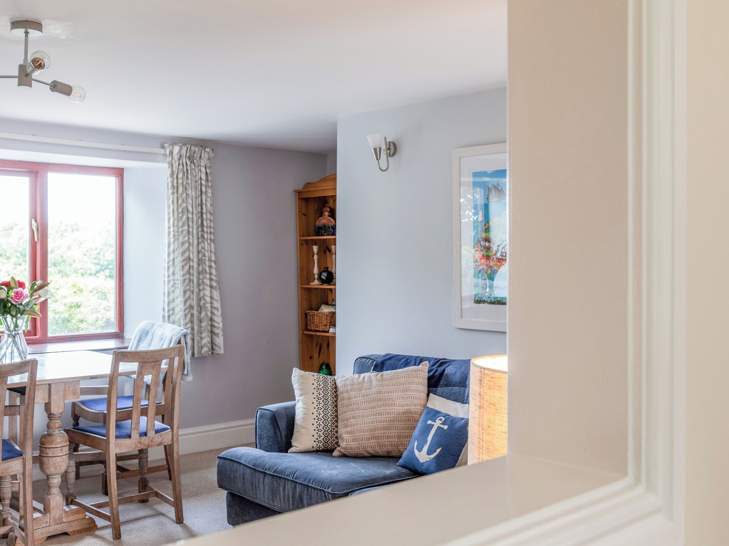 A cozy living room with a blue sofa, wooden dining table, and chairs. There is a bookshelf and a framed picture on the wall.