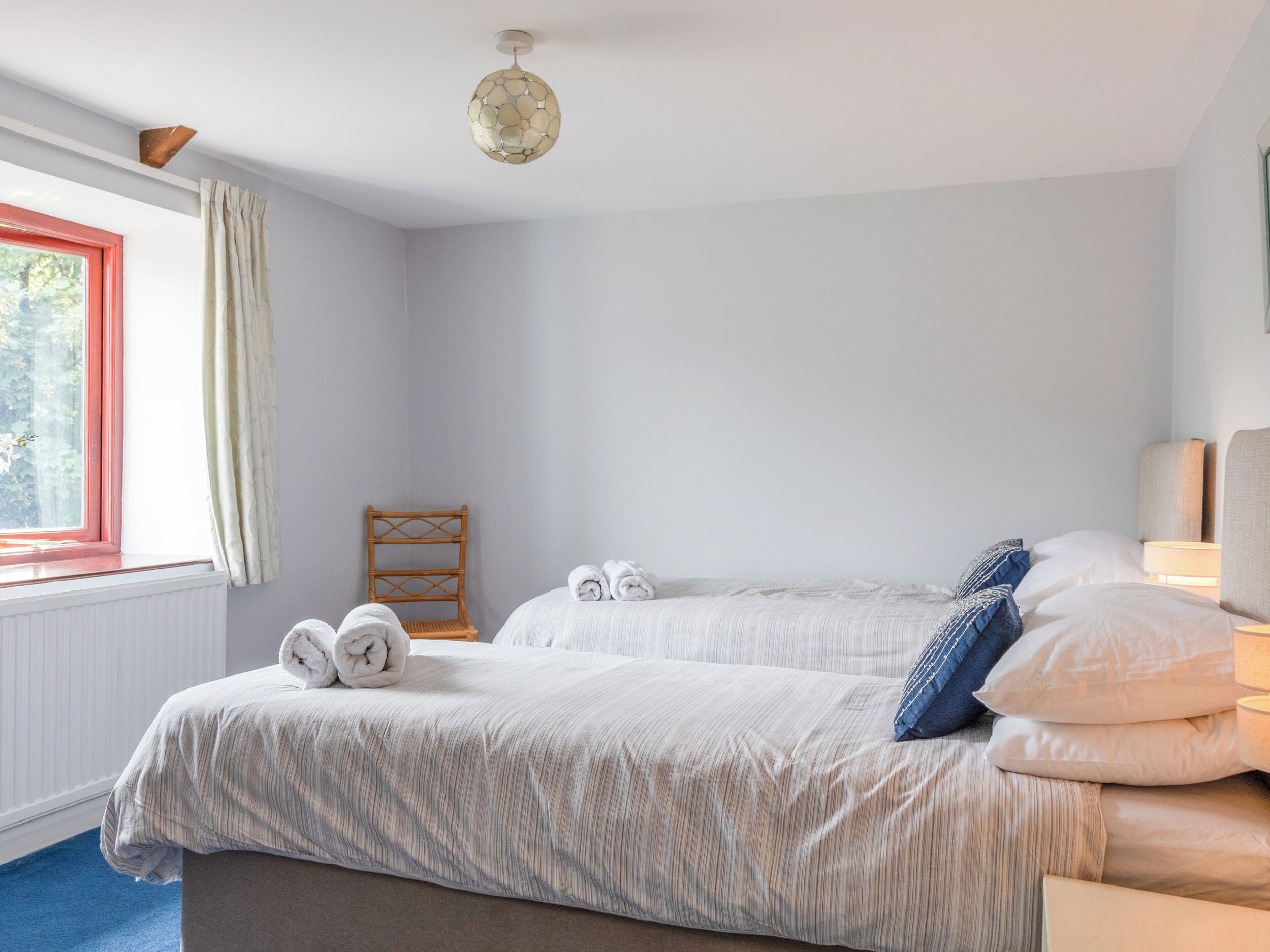 A bedroom with two beds, a wooden chair, a window with curtains, and a ceiling light fixture.