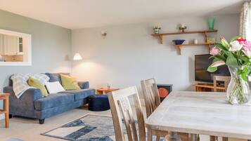 A living room with a sofa, coffee table, and dining table. There is a TV and a vase with flowers on the table.