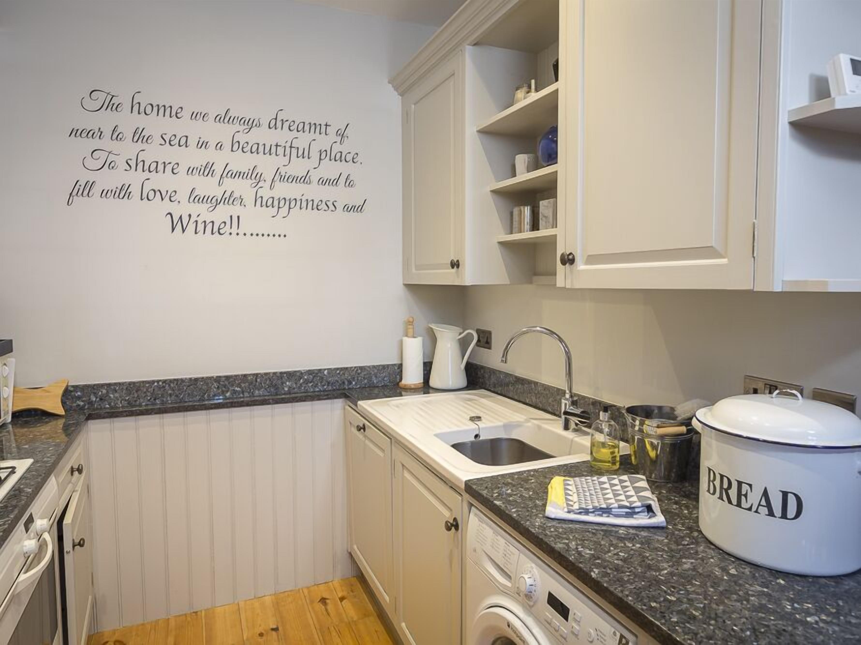 Une cuisine avec des meubles blancs, un plan de travail en granit, un évier, un récipient à pain et un mur orné d’une citation inspirante.