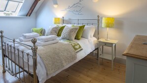 A bedroom with a sloped ceiling, a bed with white bedding and green pillows, two bedside lamps, a small bedside table, and a window with a view of the sky. - Rosemary Cottage (Kingsbridge)