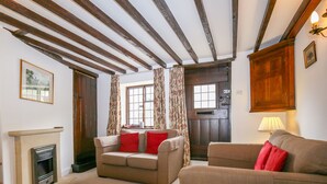 Una sala de estar con chimenea, dos sofás con cojines rojos, una puerta de madera y una ventana con cortinas.