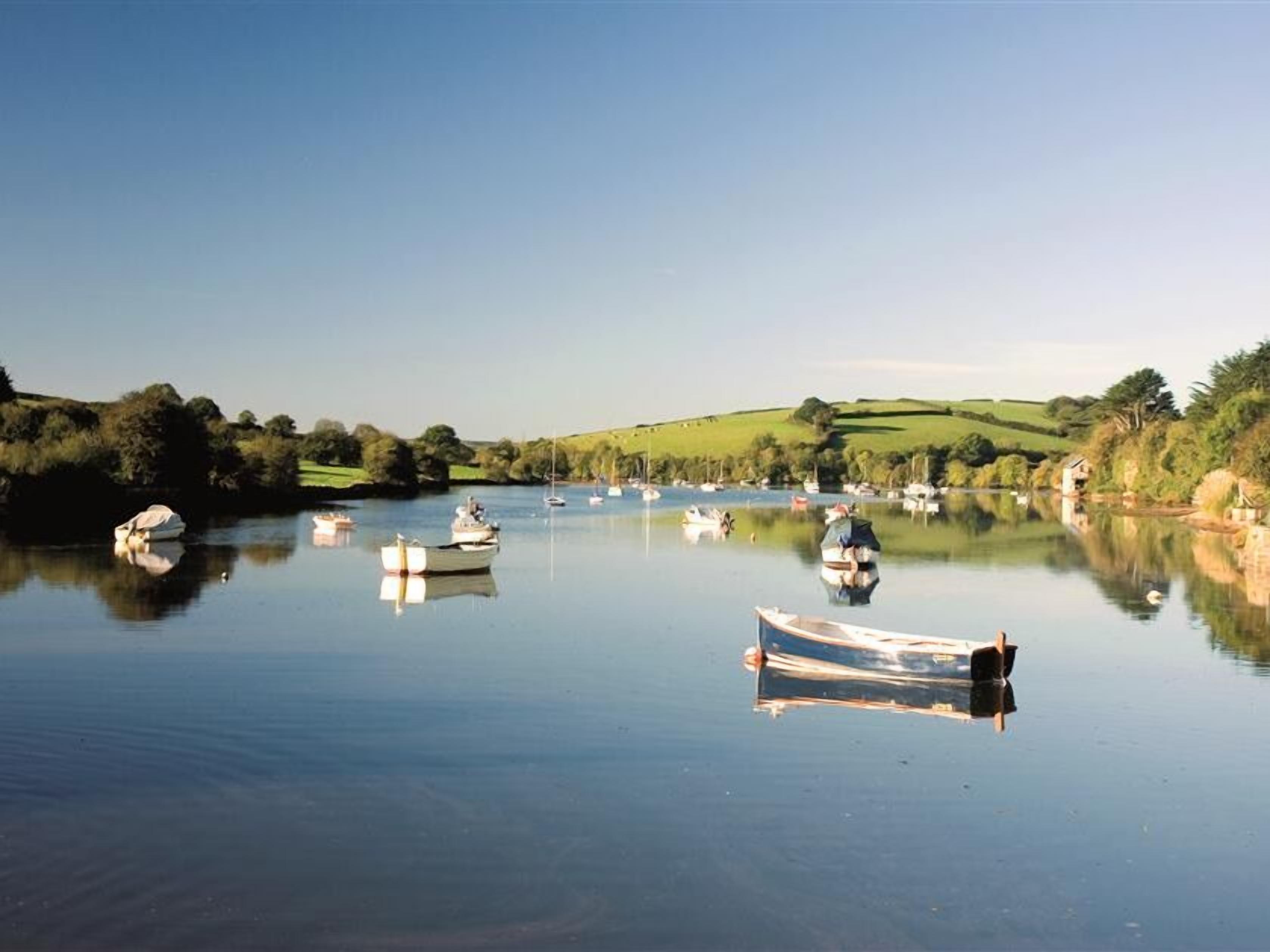 A calm river with several boats anchored, surrounded by green hills and a clear sky.