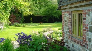 A stone building with a window, surrounded by a lush garden with various plants and a dog in the background.
