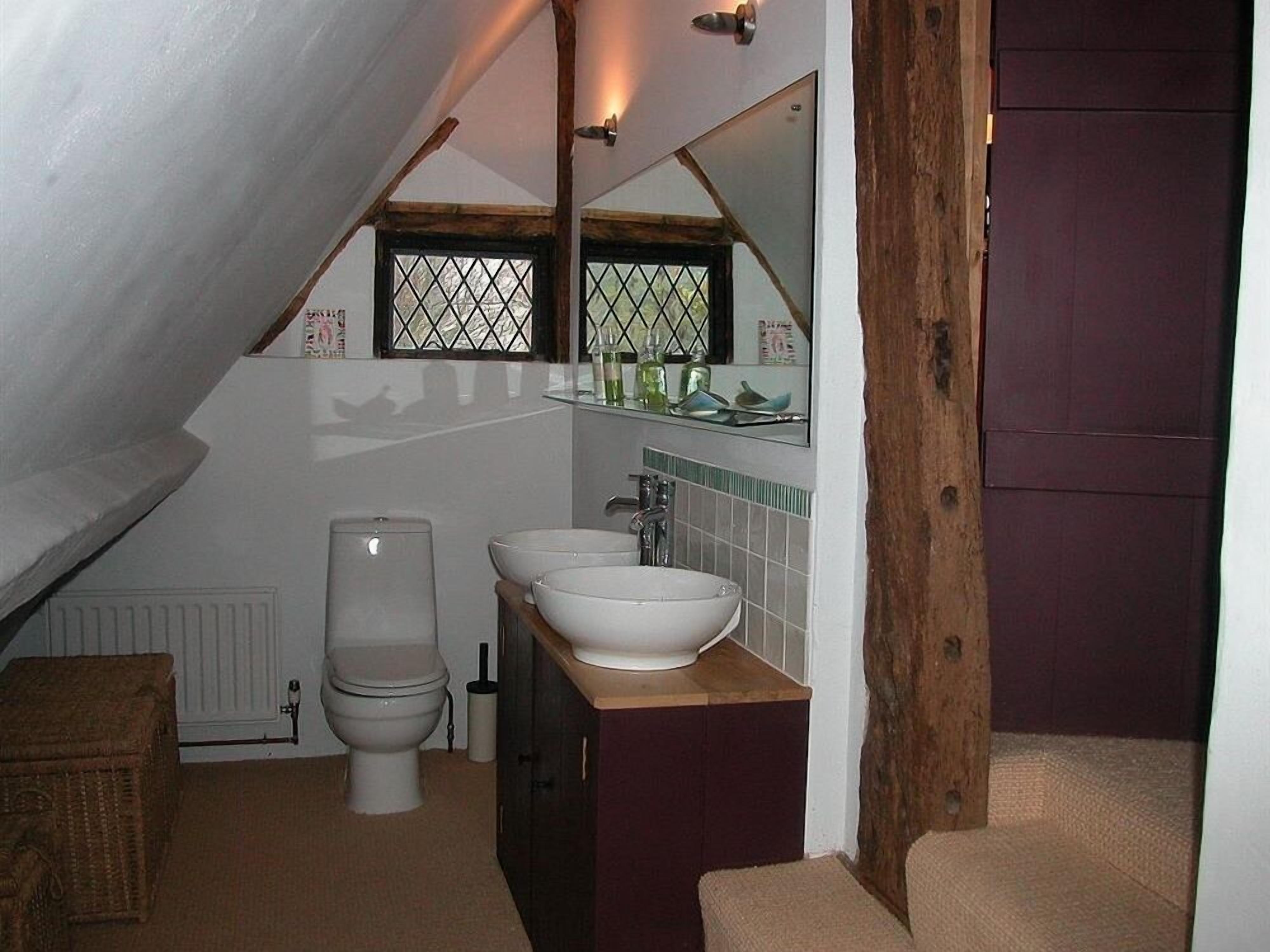 A bathroom with a sloped ceiling, a toilet, two sinks, a mirror, and a wooden door.