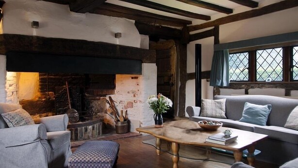 A cozy living room with exposed wooden beams, a stone fireplace, and a comfortable seating area with a sofa and armchairs.