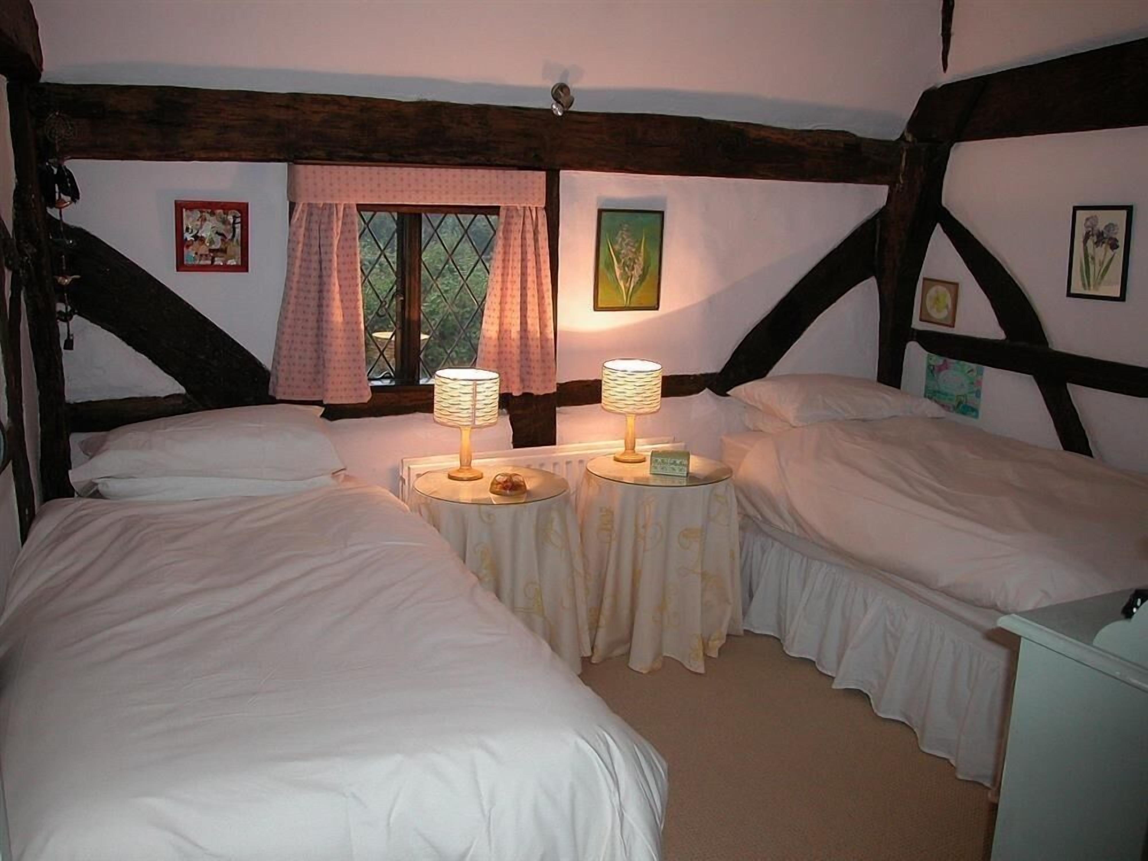 Una habitación con dos camas, cada una con mesita de noche y lámpara, una ventana con cortinas y cuadros en las paredes.