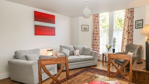A living room with a grey sofa, a wooden coffee table, and a red bowl. There is a window with floral curtains and a plant on the windowsill. - The Haven (Buxton)