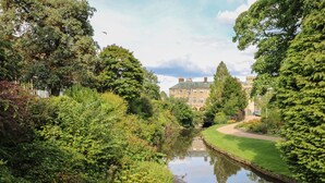 A serene riverside view with lush greenery and a historic building in the background. - The Haven (Buxton)