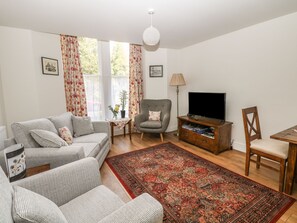 A living room with a grey sofa, a small table with a lamp, a TV stand, and a patterned rug. - The Haven (Buxton)