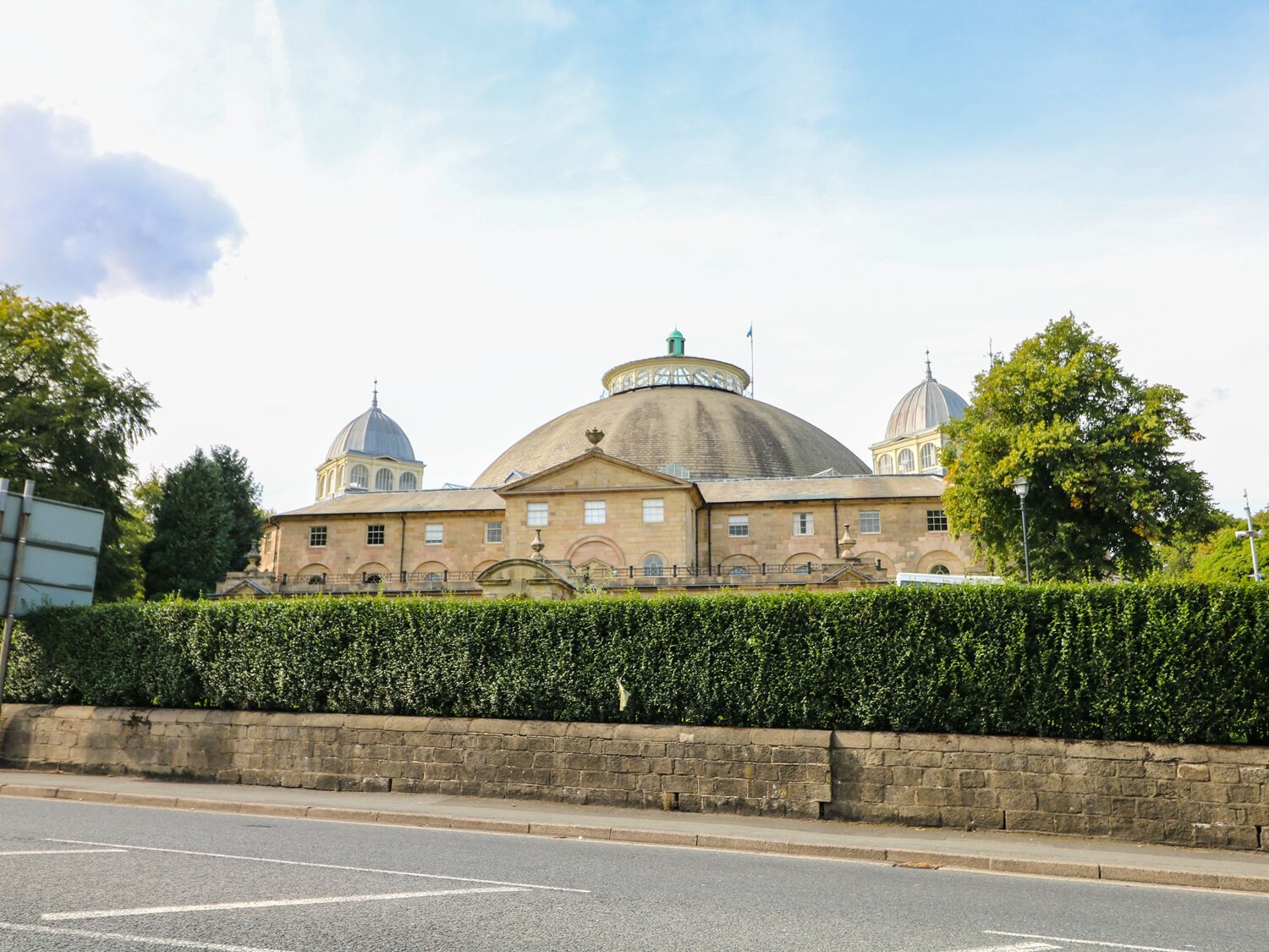 A historic building with a large central dome and smaller domes on either side, surrounded by a well-maintained hedge and a stone wall.