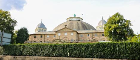 A historic building with a large central dome and smaller domes on either side, surrounded by a well-maintained hedge and a stone wall.