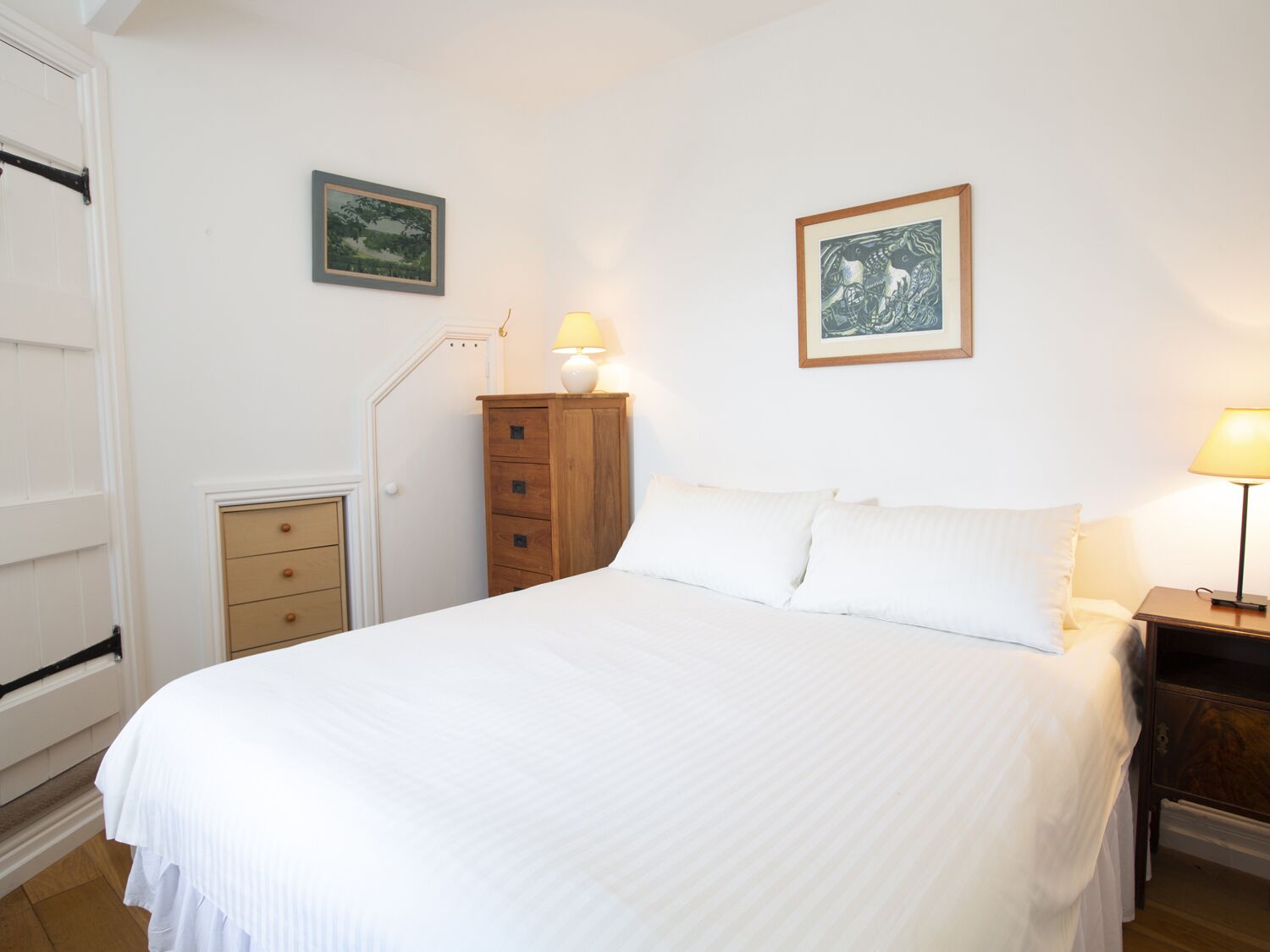 A bedroom with a large bed, a wooden dresser, two wall-mounted lamps, and two framed pictures.
