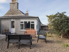 A stone cottage with a chimney, a wooden bench, and a small garden area with gravel and a tree.