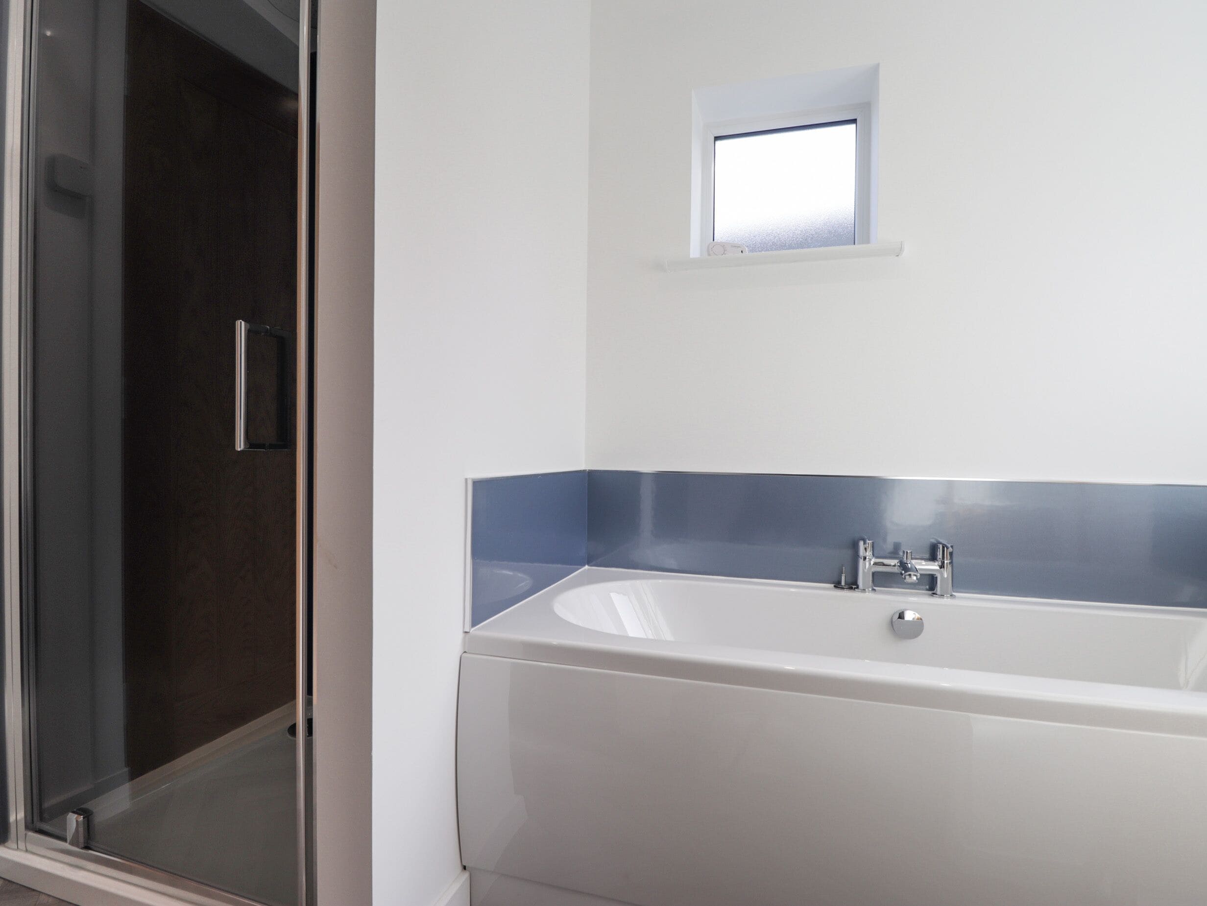 A bathroom with a glass shower enclosure, a white bathtub with a blue accent wall, and a small window above the bathtub.