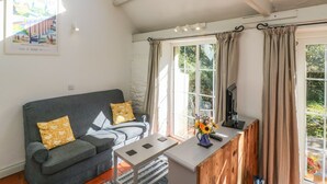 A cozy living room with a grey sofa, a small table, and a wooden cabinet. There is a skylight above the sofa and a large window with curtains. - The Daffodil Shed (Yelverton)