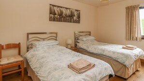 Une chambre avec deux lits simples, une chaise en bois, une table de chevet avec une lampe, une fenêtre avec des rideaux et une œuvre d’art accrochée au mur.
