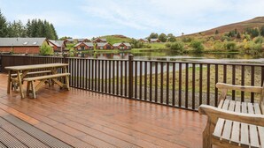 Une terrasse en bois avec des tables de pique-nique et une vue sur un lac et des bâtiments en arrière-plan.
