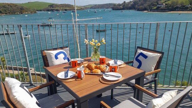 Mesa de madera dispuesta con platos, cubiertos y tazas anaranjadas, con vista a una bahía con barcos.
