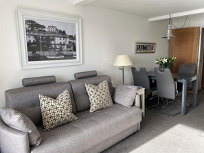 A modern living room with a grey sofa, patterned cushions, a dining table with chairs, a lamp, and a framed black and white photo on the wall. - 35 The Salcombe (Salcombe)