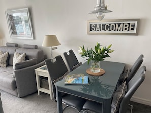 A modern living room with a dining table, chairs, a lamp, a framed sign, and a vase with flowers. - 35 The Salcombe (Salcombe)
