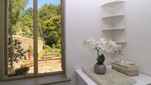 A room with a window overlooking a garden, a white shelf, and a vase with white flowers. - Garden Cottage (Bampton)