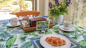 A breakfast setup with a croissant, coffee cups, a basket of chocolate cookies, and a welcome pack on a table with a tropical leaf tablecloth. - Garden Cottage (Bampton)