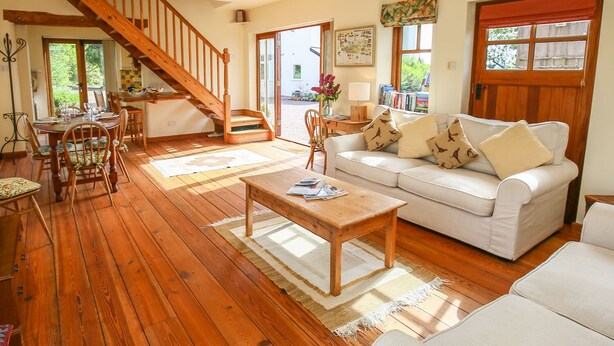 A spacious living room with a wooden staircase, a white sofa, a coffee table, and a dining area with wooden chairs and a table. - The Coach House (Exmouth)