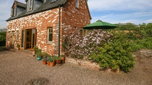A two-story brick house with a sloped roof, a front door, and a small porch area with potted plants. - The Coach House (Exmouth)