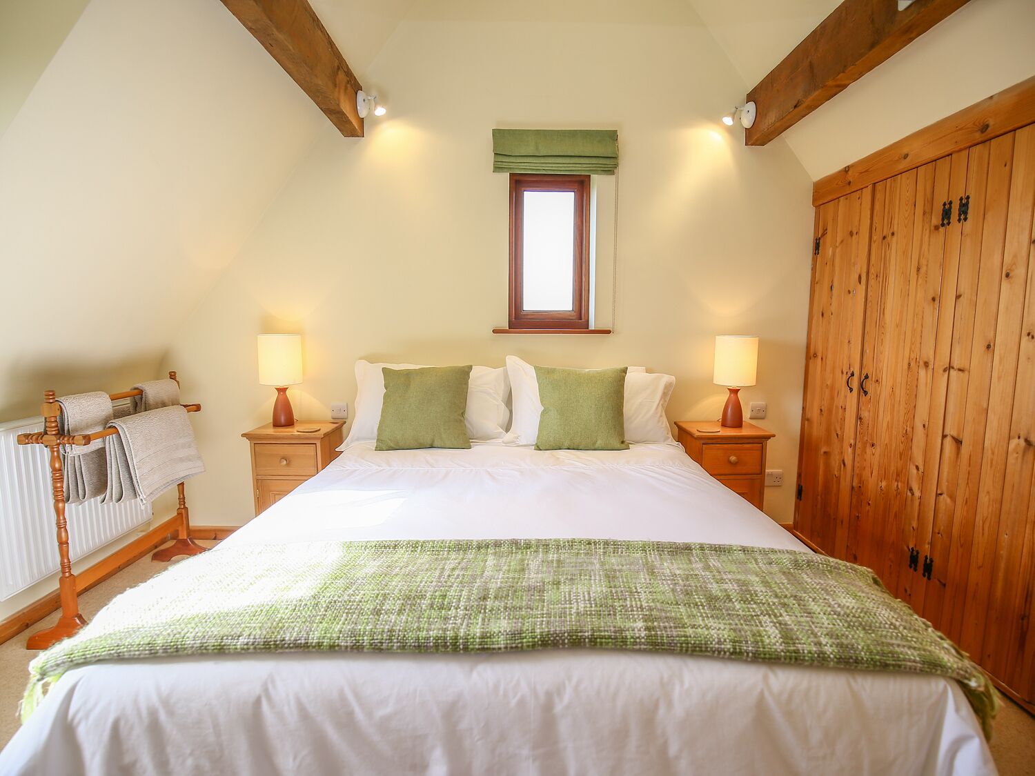 A bedroom with a large bed, two bedside tables with lamps, a wooden wardrobe, and a window with a green valance.
