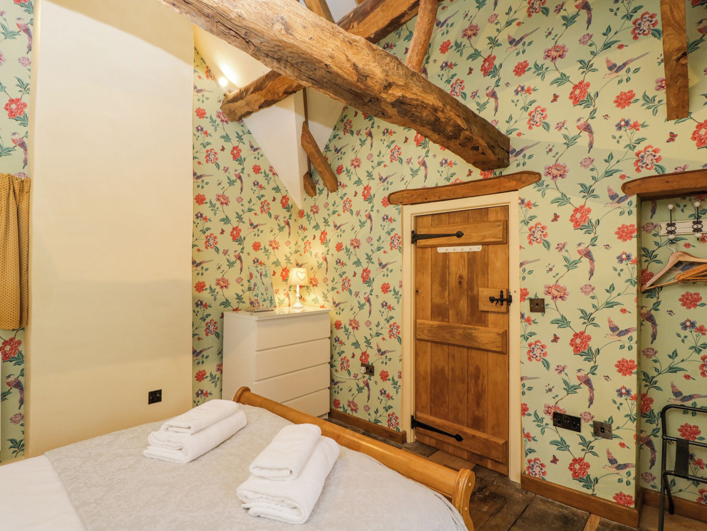 Un dormitorio con cama de madera, una mesita de noche con lámpara y papel tapiz con motivos florales.