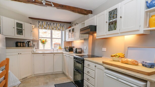 A compact kitchen with white cabinets, a Bosch dishwasher, a wooden countertop, and a window with a geometric curtain. - Godscroft Hall (Frodsham)