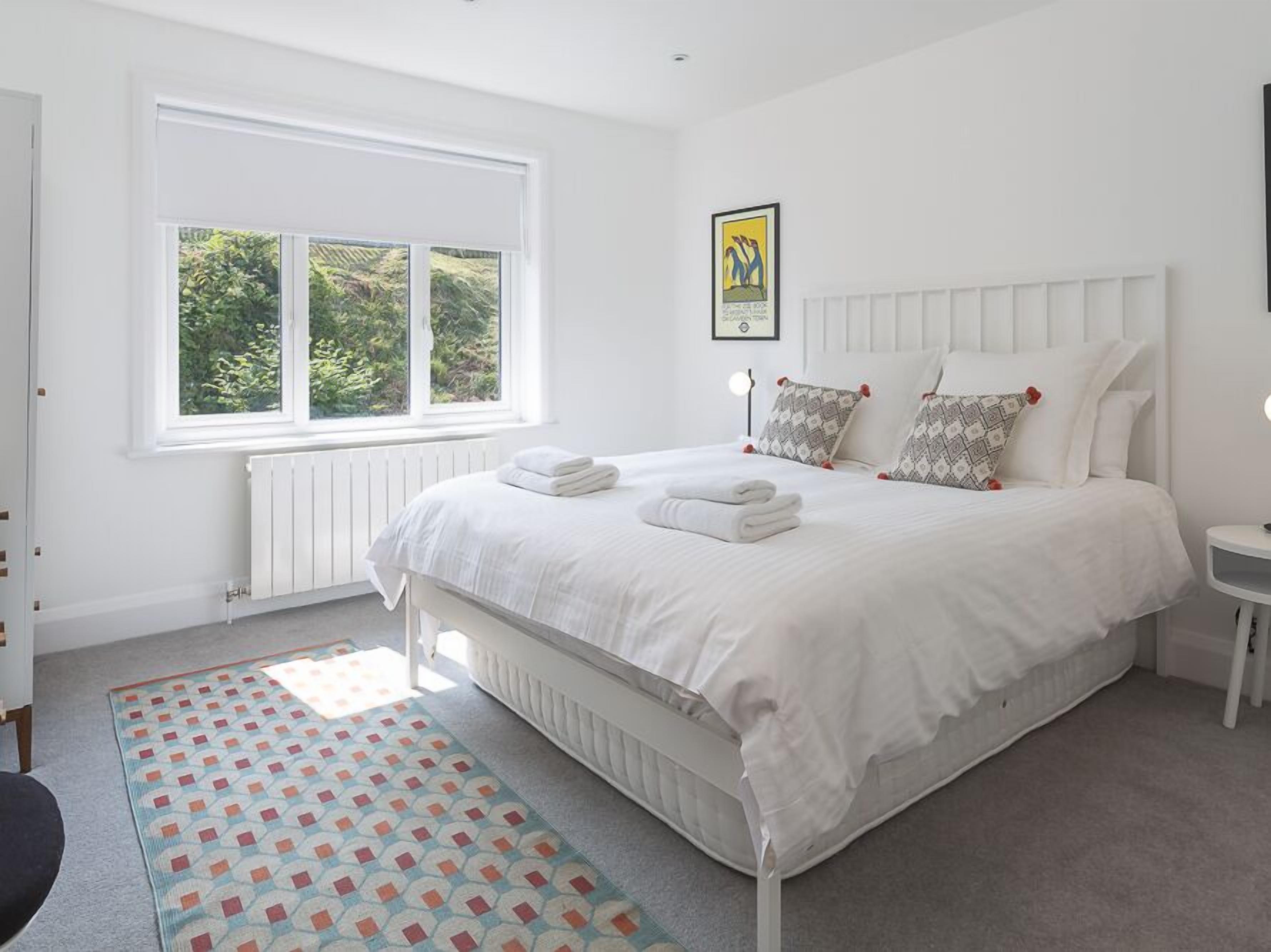 A neatly arranged bedroom with a large bed, a window with a view of greenery, and a patterned rug.