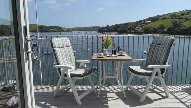 A balcony with two white chairs and a table, overlooking a body of water with boats and greenery in the background. - 36 The Salcombe (Salcombe)