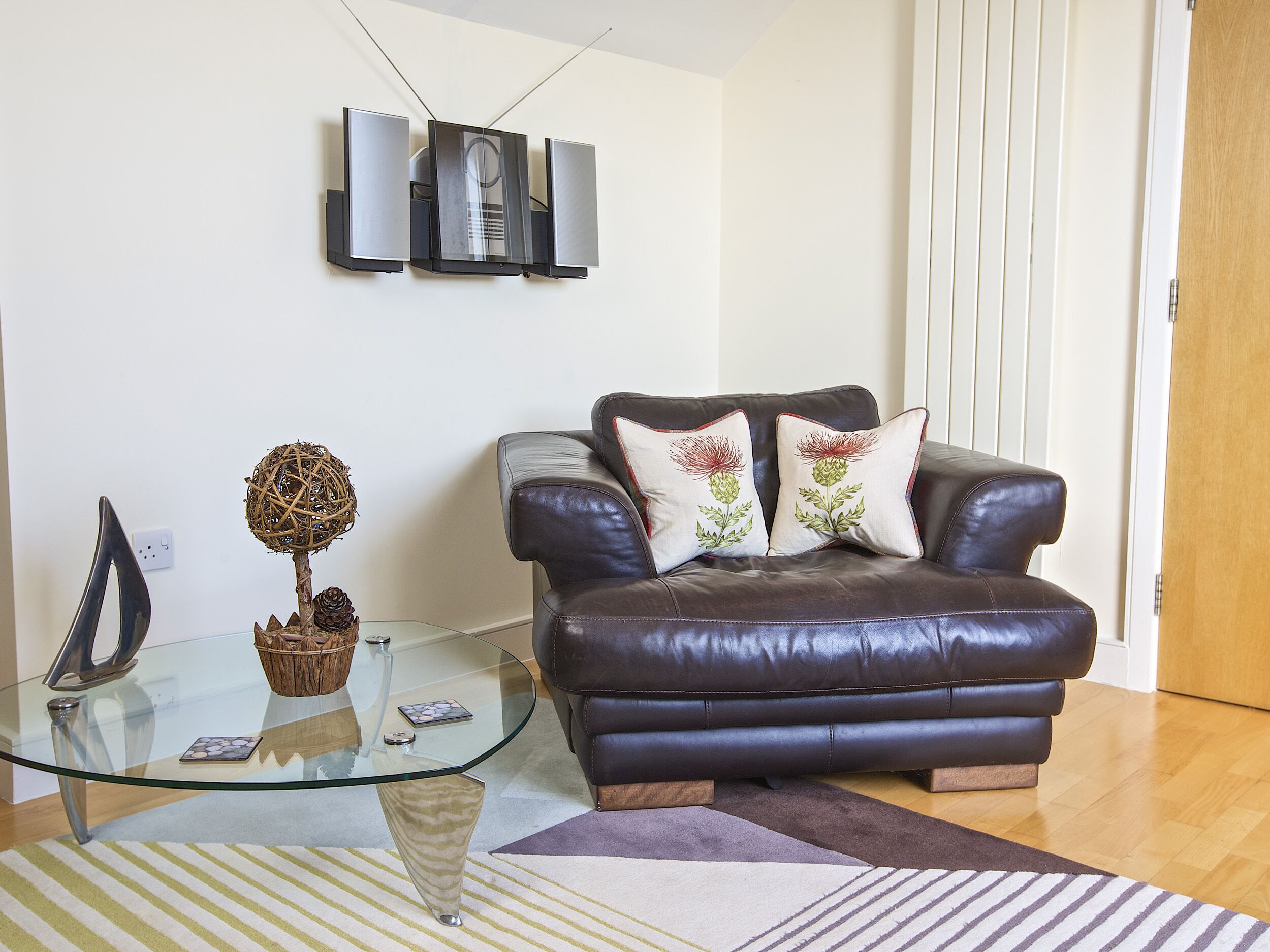Una sala de estar con una mesa de centro de vidrio, un sofá de cuero marrón, almohadones decorativos, un televisor montado en la pared y una bola de mimbre sobre una mesa de vidrio.