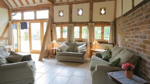 A cozy room with a high ceiling and exposed wooden beams, furnished with two sofas and a small table, and large windows allowing natural light. - Whites Farm Barn (Dymock)