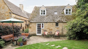 A stone cottage with a tiled roof, a wooden bench, a table with chairs, and a green umbrella. - Hadcroft Cottage (Moreton-in-Marsh)