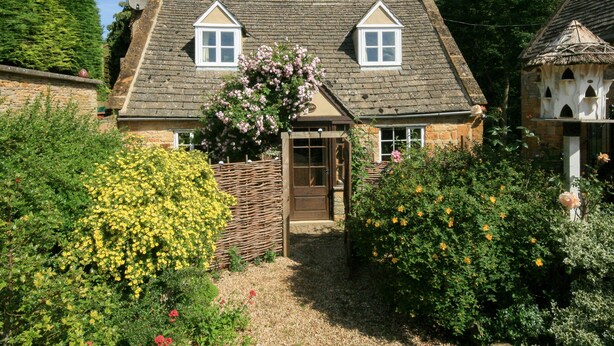 A traditional stone house with a thatched roof, a wooden gate, and a garden with flowering plants. - Hadcroft Cottage (Moreton-in-Marsh)