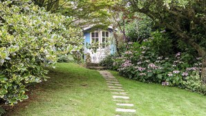 A garden pathway leading to a blue house with a white door, surrounded by lush greenery and flowering plants. - Hadcroft Cottage (Moreton-in-Marsh)