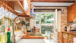 A kitchen with wooden cabinets, a sink, and a window overlooking a garden. - Hadcroft Cottage (Moreton-in-Marsh)