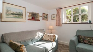 A living room with a sofa, a chair, a lamp, a window with curtains, and framed pictures on the wall. - Mayfly Cottage (Cirencester)