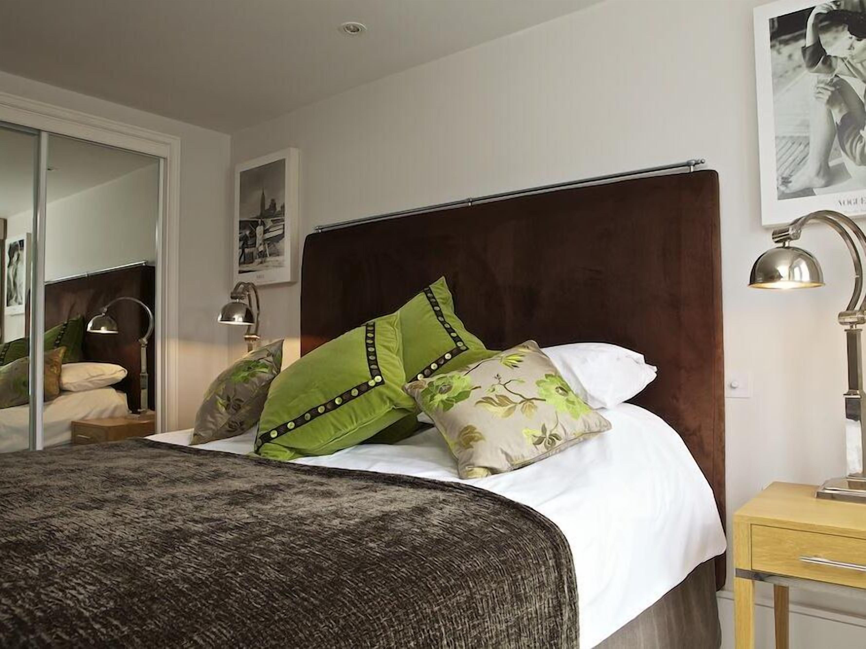 Een netjes opgemaakt bed met groene en bloemenkussens, een donkere hoofdbord en een nachtkastje met een lamp.