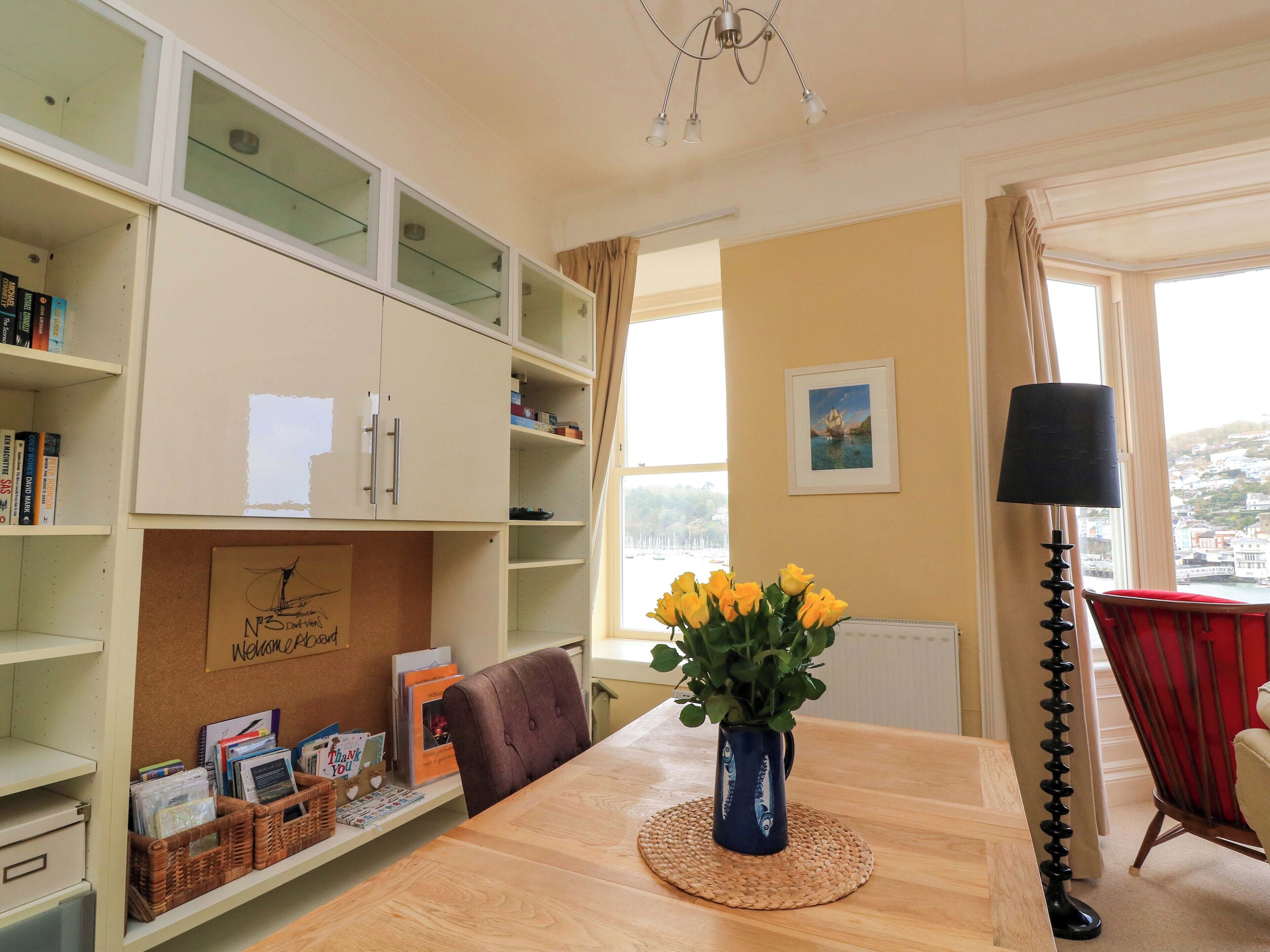 Een kamer met een houten tafel, een vaas met gele bloemen, een rode stoel, een zwarte vloerlamp en uitzicht op een stad door grote ramen.